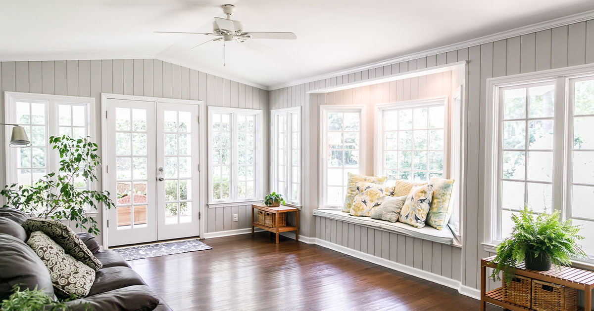 Large and open living room den sun room with windows on two sides and lots of natural light flowing in. There is a window seat on one side and a leather couch and plant on the other.