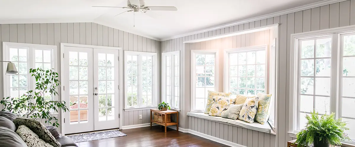 Large and open living room den sun room with windows on two sides and lots of natural light flowing in. There is a window seat on one side and a leather couch and plant on the other.