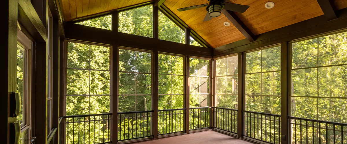 Spacious screened porch with wooden floors, big fibrex windows and ceiling fan amidst greenery.