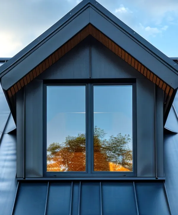 Three dormer windows on a modern house roof, with reflections of trees in the glass and a blue sky with clouds above