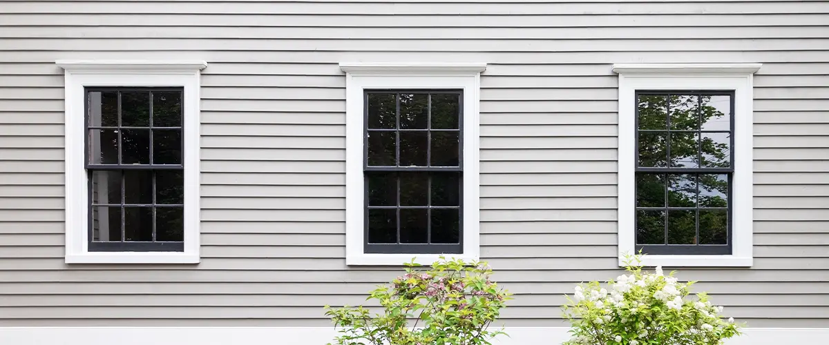 Exterior view of a home with three single-hung windows framed by gray siding and shrubs