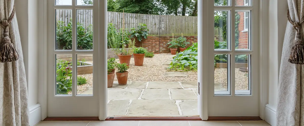 View of a backyard garden through elegant French doors with potted plants and a stone pathway.