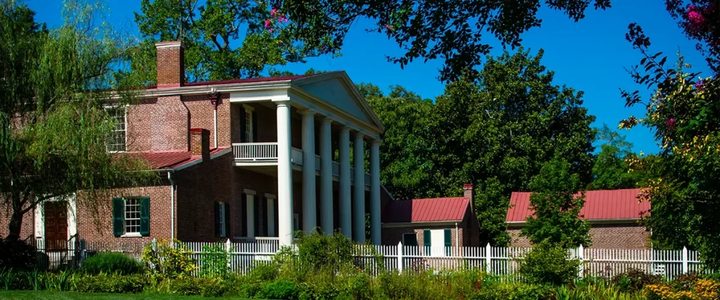 Classic brick plantation house with white columns, red roof, and lush green garden under blue sky