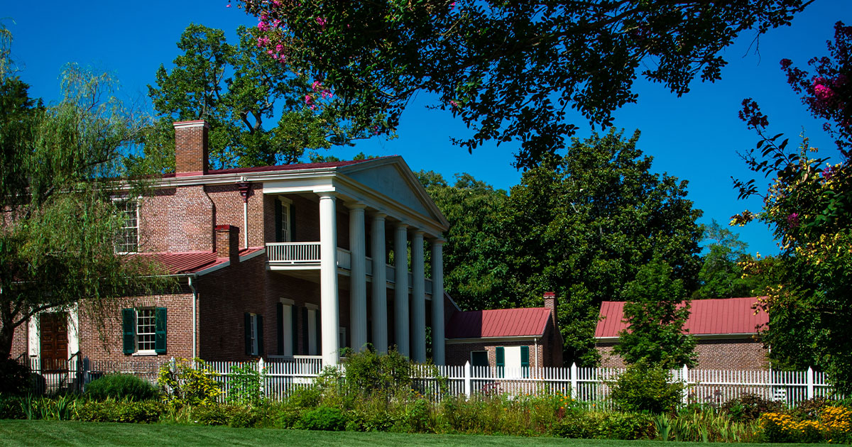 Classic brick plantation house with white columns, red roof, and lush green garden under blue sky