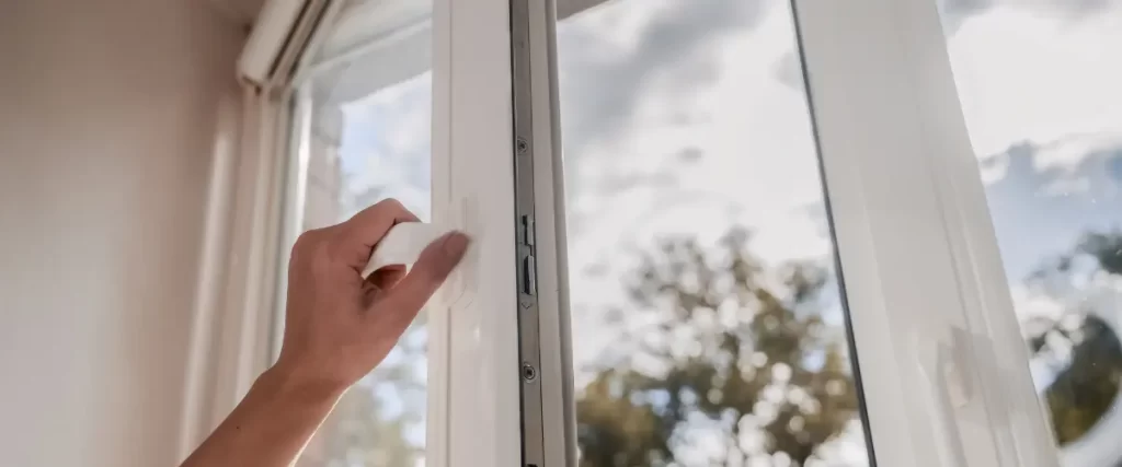 A hand opening a white window, with a blurred outdoor view of trees and sky in the background.
