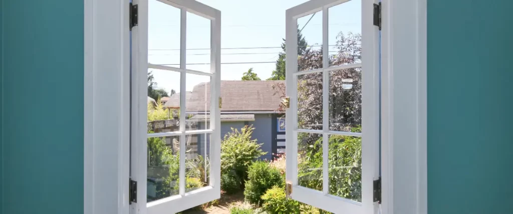 Open white-framed windows overlooking a garden with shrubs and a neighboring house in the background.