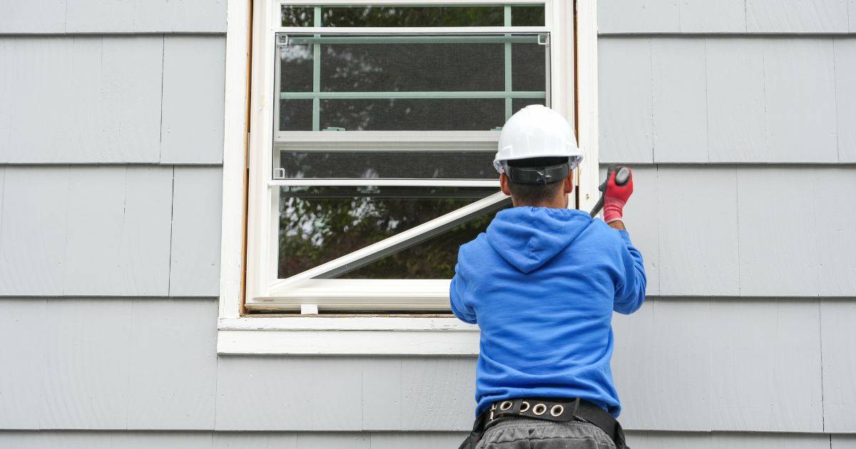 Two workers in blue hoodies and white helmets installing or adjusting a window frame on the exterior of a house.
