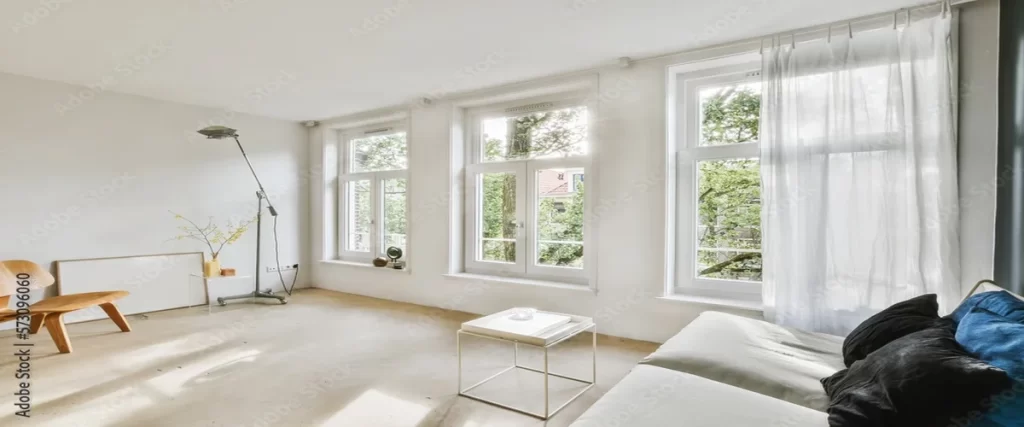 A modern living room with large white-framed windows, allowing abundant natural light and views of green trees outside. White sheer curtains hang on the right