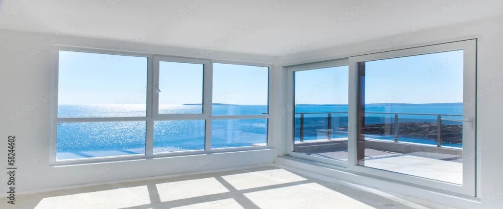 A minimalist room with large white-framed windows and a sliding glass door, offering expansive, sparkling ocean views and bright natural light