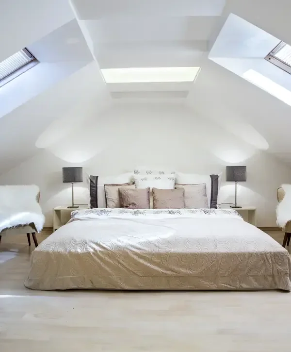 Modern attic bedroom with white sloped walls and ceiling, featuring a large bed with neutral beige bedding, two bedside tables with dark lamps, and multiple skylight windows for natural light.