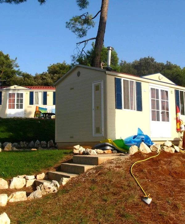Modern prefabricated home with cream siding, blue shutters, and stone steps leading up a hillside, set in a wooded area with pine trees and green landscaping.