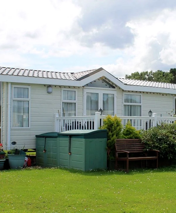 Exterior of a white prefabricated home with a pitched roof and large windows, showcasing two green outdoor storage units and a single wooden bench on a well-kept lawn under a cloudy sky.