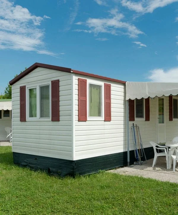 White home exterior with horizontal siding, red shutters, and a dark red roof trim, located on a green lawn with a small patio and a temporary white awning attached to the side.