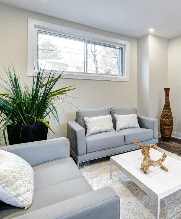 a basement living room with a gray sofa, a gray accent chair, a white coffee table, and large windows