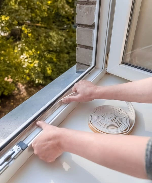Hands carefully pressing a self-adhesive foam weatherstripping roll into the track of an open white window frame to ensure energy-efficient windows sealing against drafts.